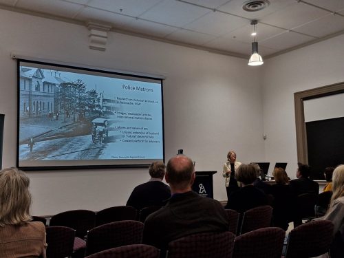 A woman presenting in front of a seated audience, the powerpoint slide next to her is on Police Matrons